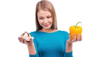 Temptation during my diet. Beautiful young woman holding a salad pepper in one hand and a cake in another  while standing against white background photo