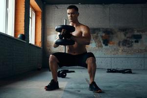 Confident young man standing in squatting position while training with dumbbell in gym photo