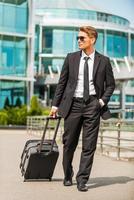 hombre de negocios en movimiento. joven hombre de negocios confiado en traje completo que lleva una maleta mientras camina al aire libre foto