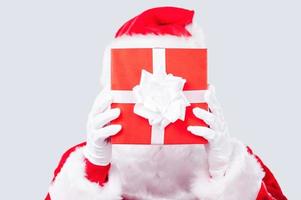 regalo de Navidad. santa claus tradicional escondiendo su rostro detrás de una caja de regalo mientras se enfrenta a un fondo gris foto