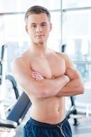 Confident strong man. Confident young muscular man keeping arms crossed and looking at camera while standing in gym photo