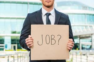 Looking for a job. Close-up of young man in formalwear holding poster with job text message while standing outdoors and against building structure photo