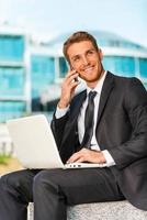 Businessman working outdoors. Handsome young man in formalwear working on laptop and talking on the mobile phone while sitting outdoors and against building structure photo