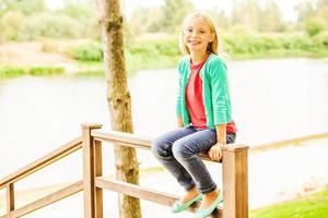 So good to do nothing. Cute little girl sitting at the wooden railing and smiling with river in the background photo