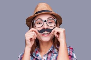 Having fun with face mustache. Cheerful young woman in funky hat holding fake mustache on her face and looking at camera while standing against grey background photo