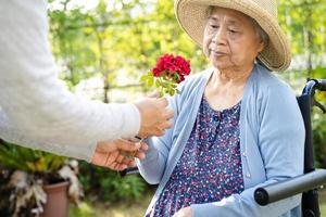 Caregiver daughter hug and help  Asian senior or elderly old lady woman holding red rose on wheelchair in park. photo