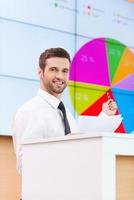 Look at the diagram Cheerful young man in formalwear standing at the tribune and pointing projection screen while making a presentation photo