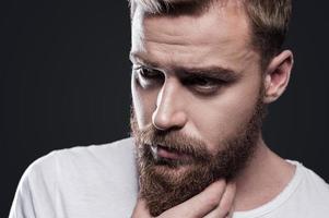 Lost in thoughts. Portrait of thoughtful young bearded man looking away and holding hand on chin while standing against grey background photo