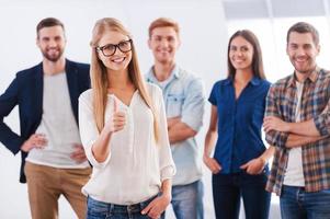 Join a successful team Beautiful young woman showing her thumb up and smiling while group of happy young people standing on background and smiling photo