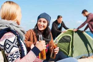 Taking time to have a rest. Two beautiful young women talking to each other and smiling while holding bottles with beer with two men setting up tent in the background
