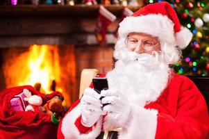 Typing message to elfs. Happy Santa Claus typing a message on the mobile phone and smiling while sitting at his chair with fireplace and Christmas Tree in the background photo