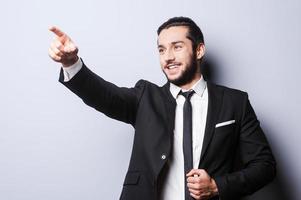 Look at that Smiling young man in formalwear pointing away while standing against grey background photo