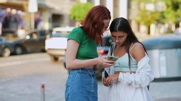 Two young women look together at phone while standing outside near a street video