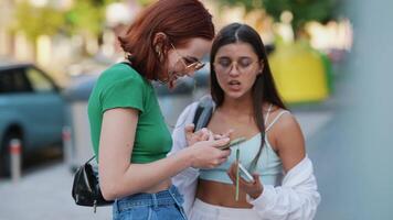 Two young women look together at phone while standing outside near a street video