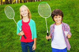 estamos listos para jugar con dos lindos niños pequeños sosteniendo raquetas de bádminton y sonriendo mientras estamos juntos en la hierba verde foto