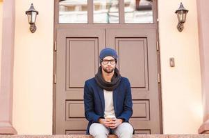 casualmente guapo. un joven apuesto con ropa informal elegante sentado frente a la entrada de la casa y sosteniendo una taza de café foto