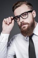He got creative mind. Handsome young bearded man in shirt and tie adjusting his eyeglasses and looking at camera while standing against grey background photo