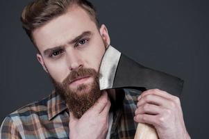 Shaving with axe. Confident young bearded man carrying a big axe on shoulder and looking at camera while standing against grey background photo