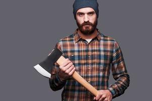 Serious lumberjack. Confident young bearded man holding a big axe and looking at camera while standing against grey background photo