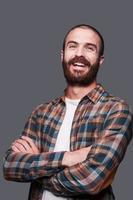 Happy bearded man. Handsome young bearded man keeping arms crossed and smiling while standing against grey background photo