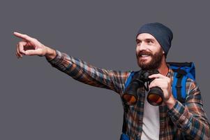 That is amazing Handsome young bearded man with backpack holding binoculars and pointing away with smile while standing against grey background photo