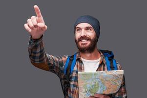 I know the right direction Happy young bearded man with backpack holding map and pointing away while standing against grey background photo