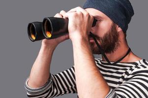 Exploring new places. Side view of confident young bearded man in striped clothing looking through binoculars  while standing against grey background photo