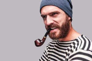 As rough as he is. Serious young bearded man in striped clothing smoking a pipe and looking away while standing against grey background photo