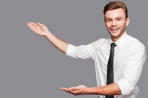 Positive selling. Confident young man in shirt looking at camera and pointing away while standing against grey background photo