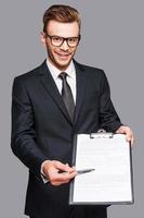 Please sign here. Portrait of confident young man in formalwear stretching out clipboard with paper and pointing it with pen while standing against grey background photo