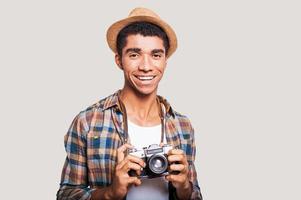 conseguir las mejores fotos. guapo joven hipster afroamericano sosteniendo la cámara y sonriendo mientras está de pie contra el fondo gris foto