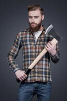 Lumberjack style. Handsome young bearded man holding a big axe and looking at camera while standing against grey background photo