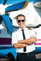 My job is my passion. Confident male pilot in uniform keeping arms crossed and smiling while standing in front of the airplane photo