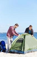 It is almost done. Two confident young men setting up a tent while standing at the riverbank photo