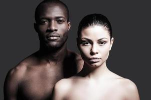 Black and white. Portrait of shirtless African man and Caucasian woman bonding to each other and looking at camera while standing against grey background photo