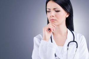 Thinking about this problem.  Female doctor in white uniform holding hand on chin while standing against grey background photo