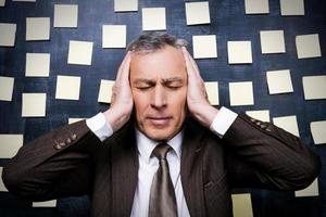 Tired from it all. Frustrated senior man in formalwear holding head in hands and keeping eyes closed while standing against blackboard with adhesive notes on it photo