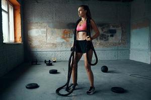 Full length of beautiful young woman carrying battle rope while standing in gym photo