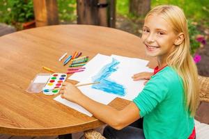 Little artist at work. Top view of cute little girl drawing something on paper and smiling while sitting at the table and outdoors photo