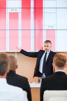 Talking about company achievements. Confident mature man in formalwear pointing projection screen with graph on it while making presentation in conference hall with people on the foreground photo