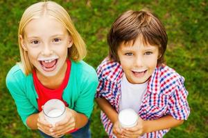 We love milk. Top view of two cute little children with milk moustaches holding glasses with milk and smiling while standing on green grass together photo