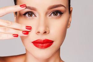 rojo clásico. primer plano de una hermosa mujer con manicura roja sosteniendo los dedos en la cara mientras está de pie contra el fondo gris foto