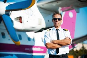 Ready for the skies. Confident male pilot in uniform keeping arms crossed while standing near airplane photo