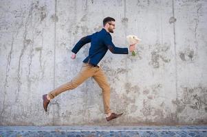 Hurrying to be in time. Side view of happy young man holding bouquet of flowers while running in front of the concrete wall photo