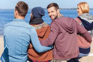 pasar un buen rato con amigos. vista trasera de cuatro jóvenes felices disfrutando del tiempo en la playa mientras un joven mira por encima del hombro y sonríe foto