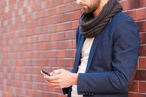 Typing message to friend. Side view of handsome young man in smart casual wear holding mobile phone while leaning at the brick wall photo