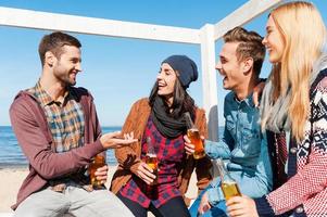 Young and carefree. Four happy friends talking to each other and smiling while sitting on the beach and drinking beer photo