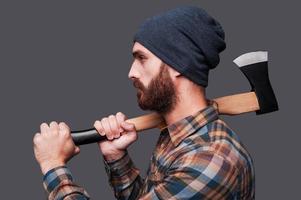 Confident lumberjack. Side view of confident young bearded man holding a big axe and looking at camera while standing against grey background photo
