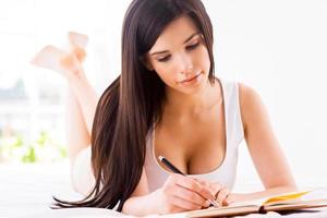 Beautiful and creative. Attractive young woman writing something in note pad while lying in bed photo