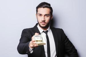 Come to work and get your first salary. Close-up of young man in formalwear stretching out money while standing against grey background photo